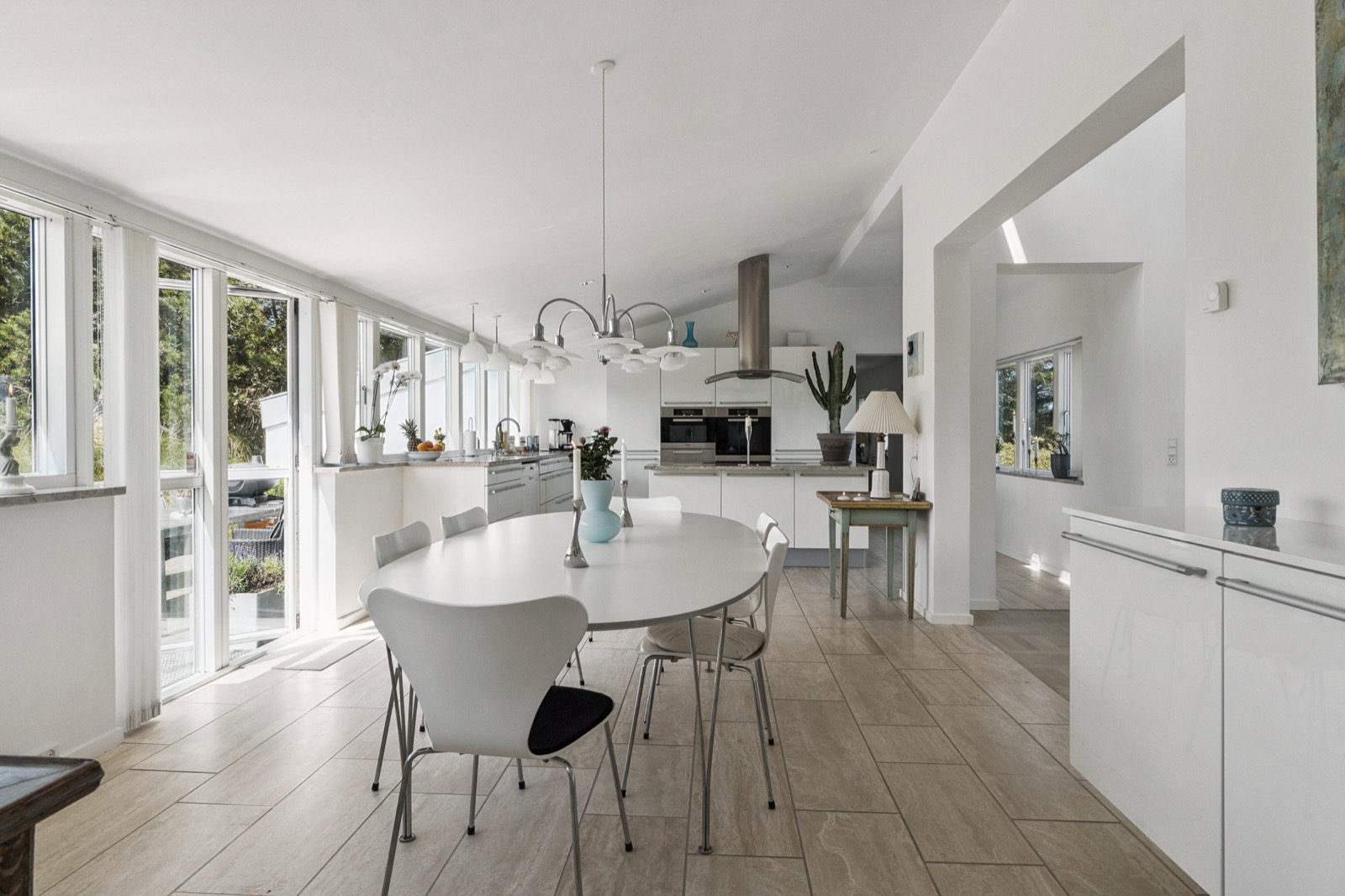 Åbent køkken-alrum med ovalt hvidt bord, lysekrone og glasdøre ud til terrasse og have.