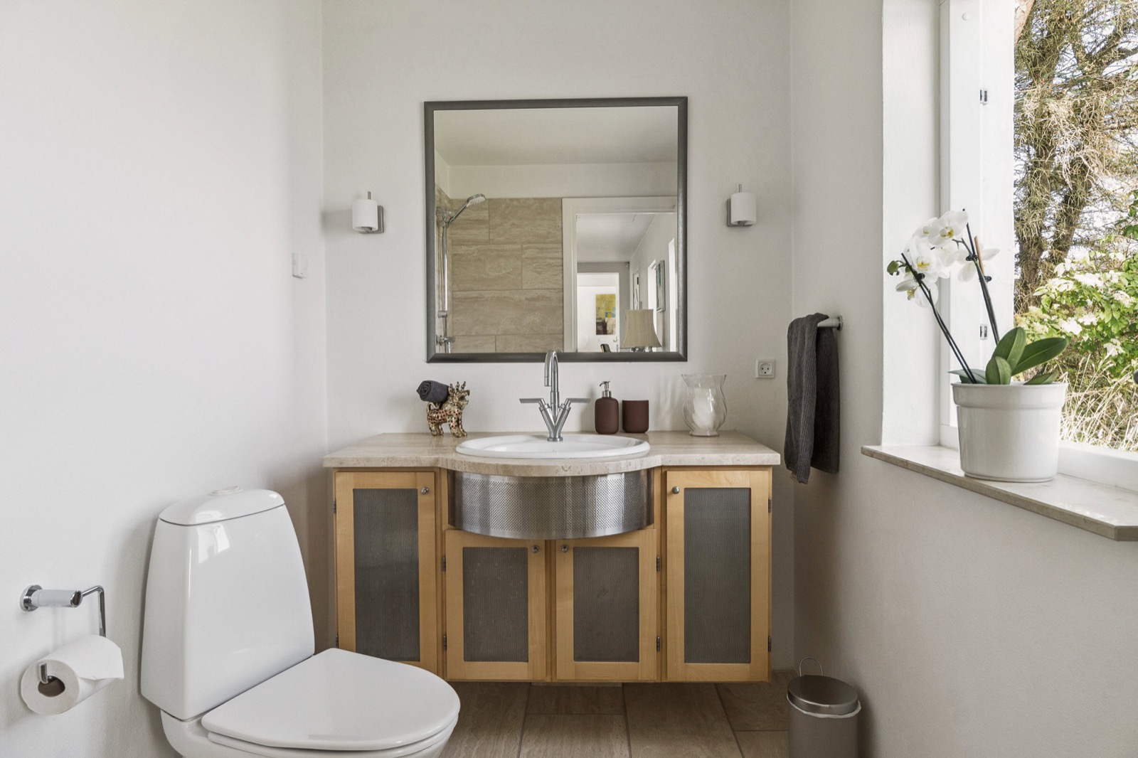 Gæstetoilet med træ- og netvaskeskab, rund stålvask og orkidé ved vinduet.