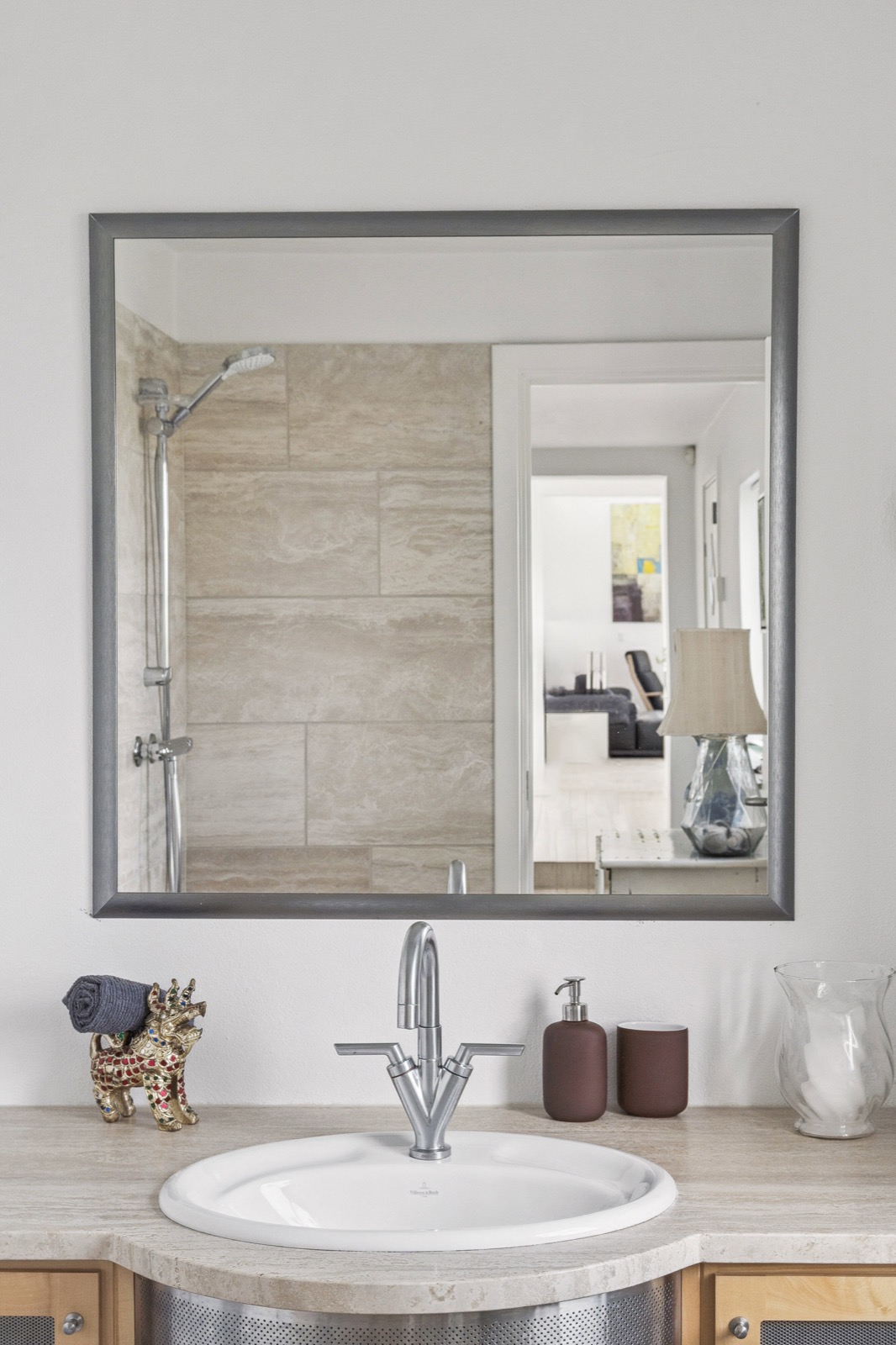 Badeværelsesmøbel med oval Villeroy & Boch-vask og stort spejl, der reflekterer brusenichen i travertin og et glimt mod stuen.