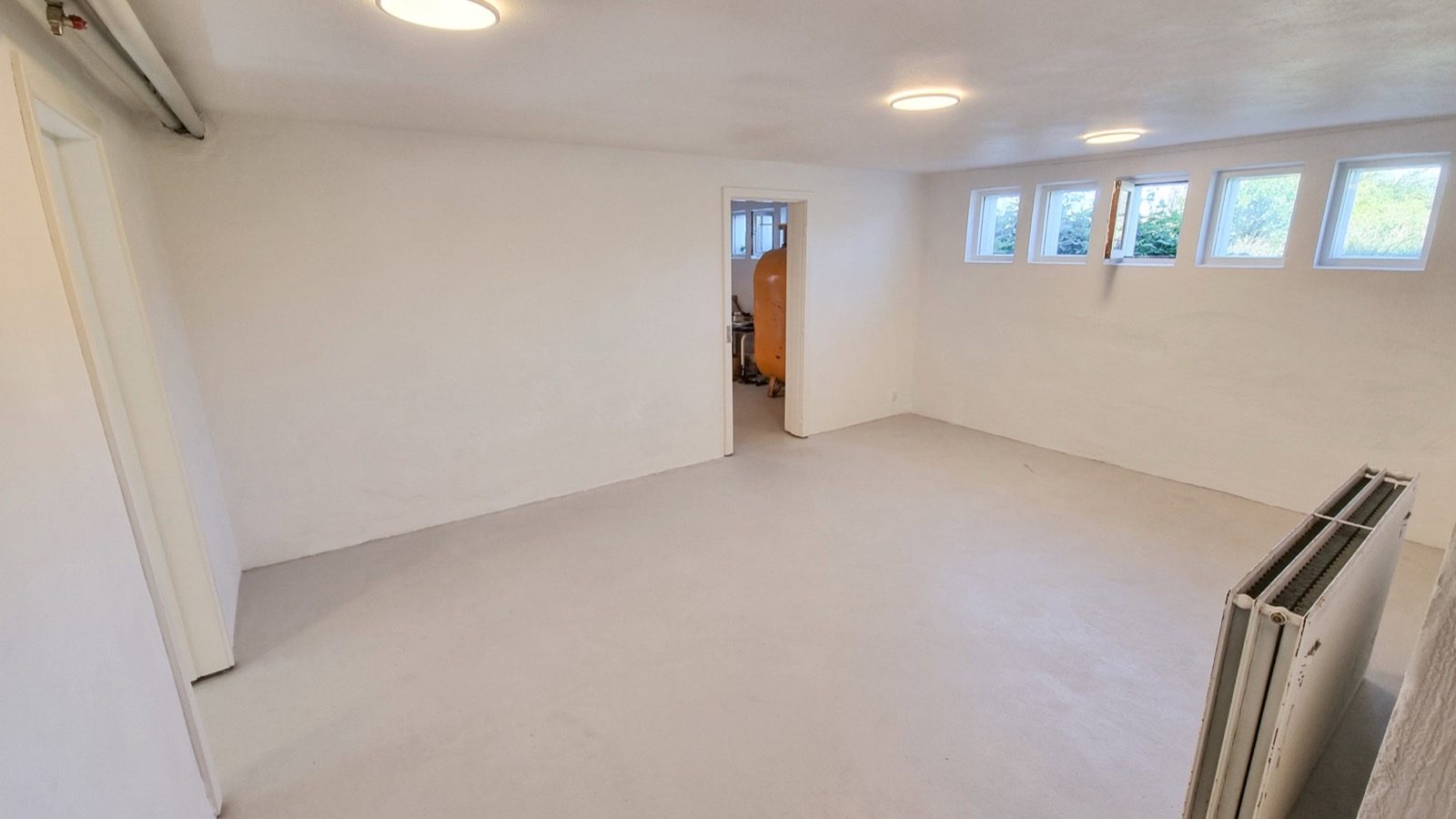 Stort råt kælderrum med betongulv, høje små vinduer, dør mod teknikrum og radiator i hjørnet.