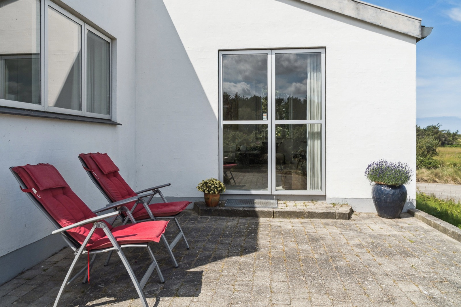 Solrig fliseterrasse med to røde liggestole ved den hvide gavl, dobbeltdøre med genskin af himmel og træer og krukker med blomster.