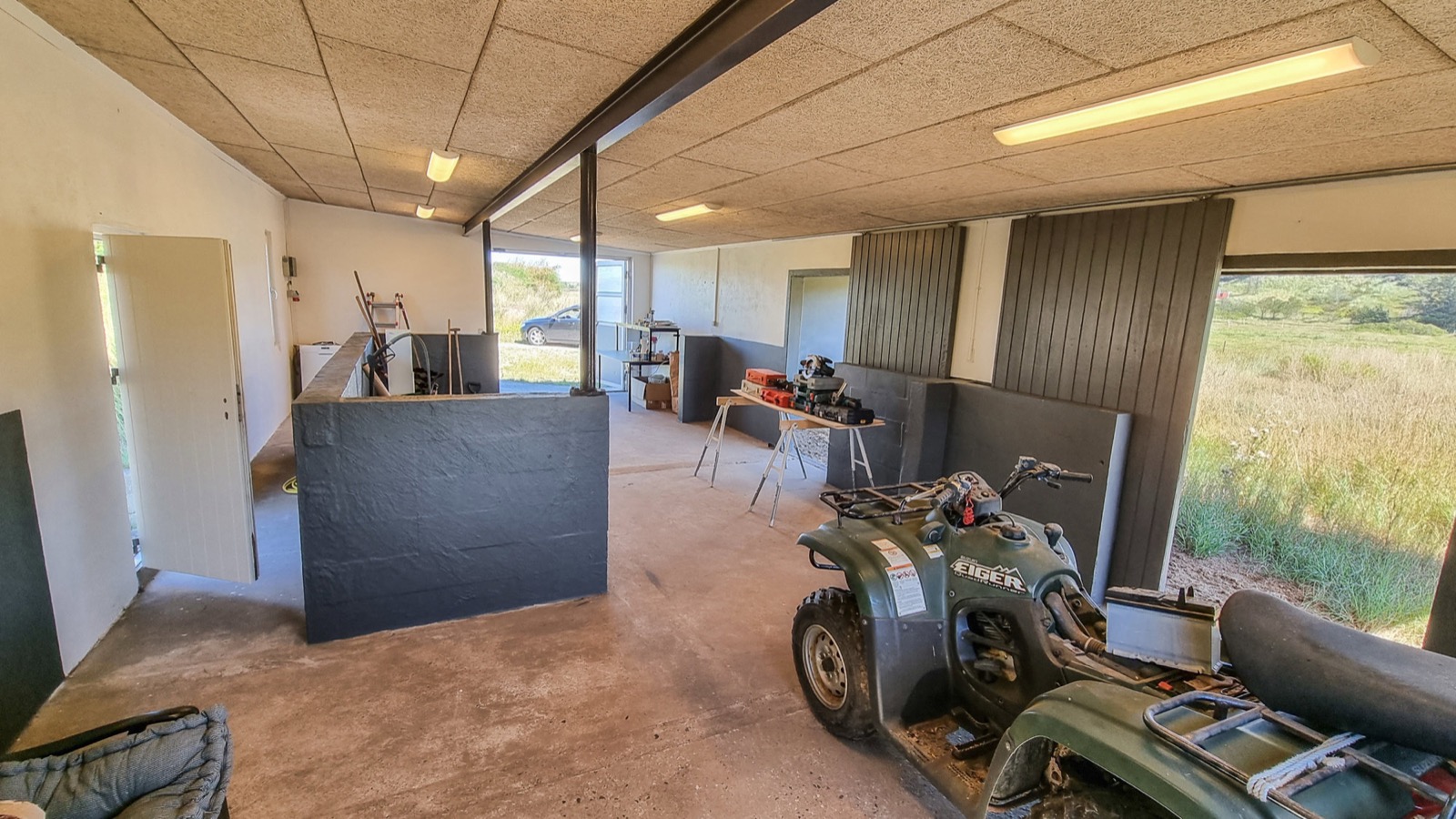 Anden vinkel af garagen og værkstedet med ATV, savbuk med elværktøj, skillevæg og åbne porte mod bil og klitlandskab.