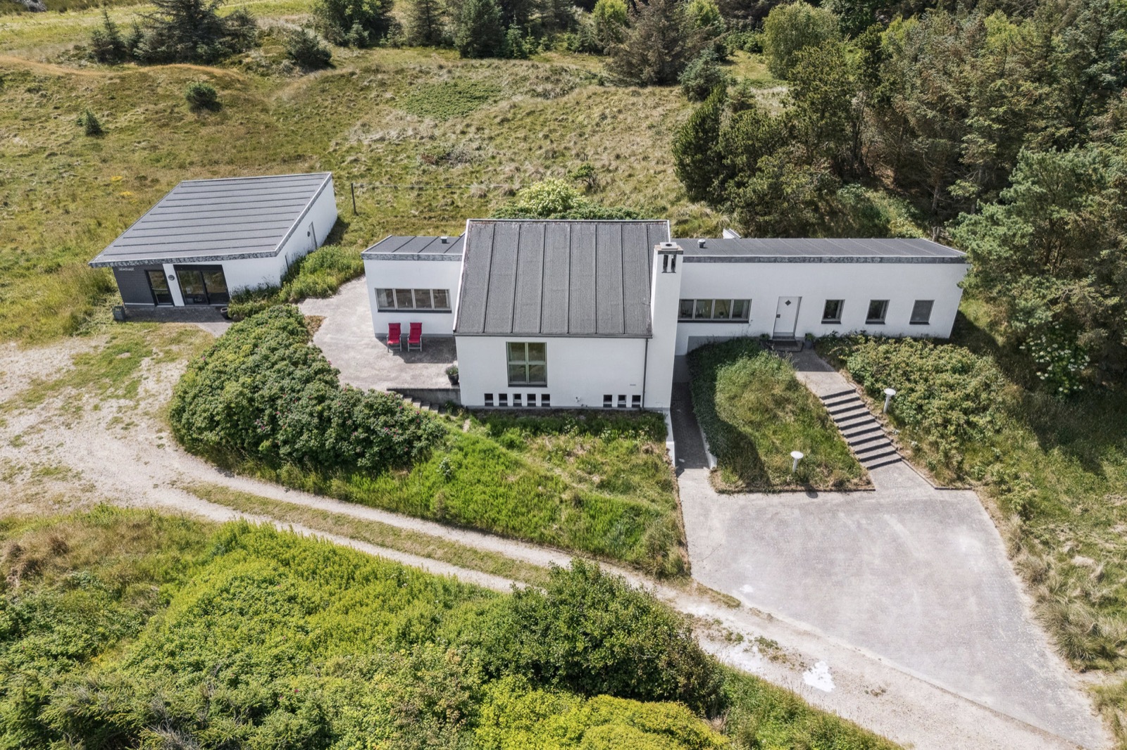 Dronefoto af hele ejendommen: hovedhus, vinklet taglinje, skorsten, fritliggende garage, grusvej, klitter og skov.
