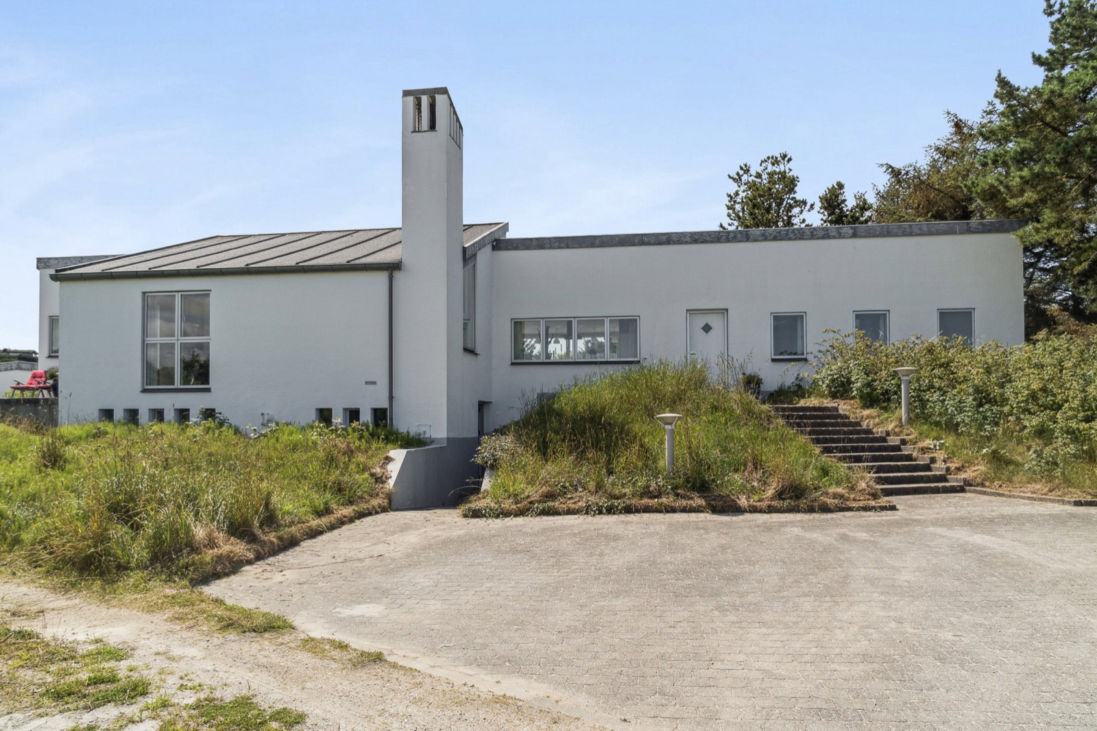 Huset set fra terræn med hvid facade, høj skorsten, indgangsdør, trappe og brolagt gårdrum.