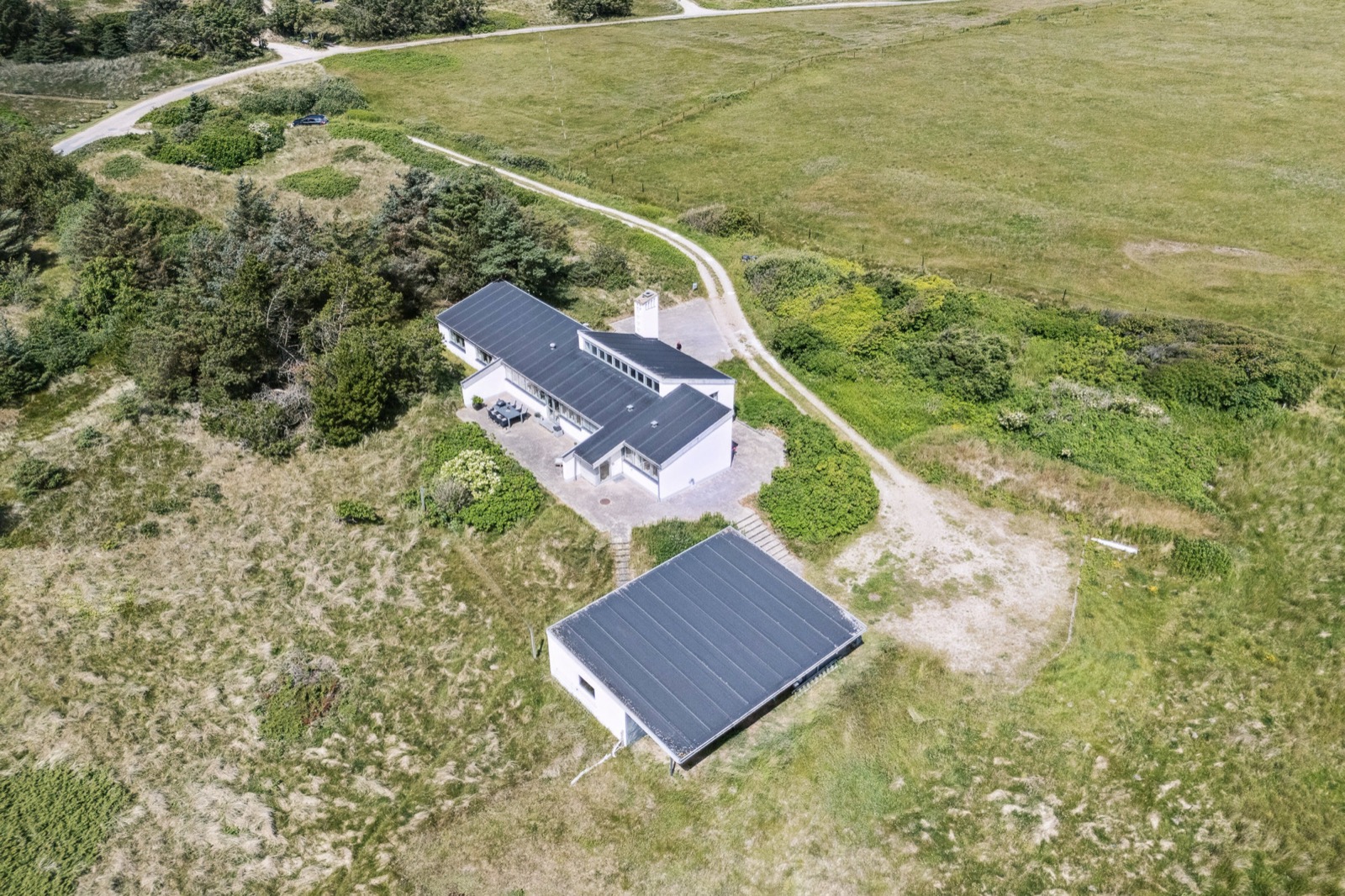 Dronefoto af hovedhuset, den fritliggende garage og grusvejen gennem klitterne.