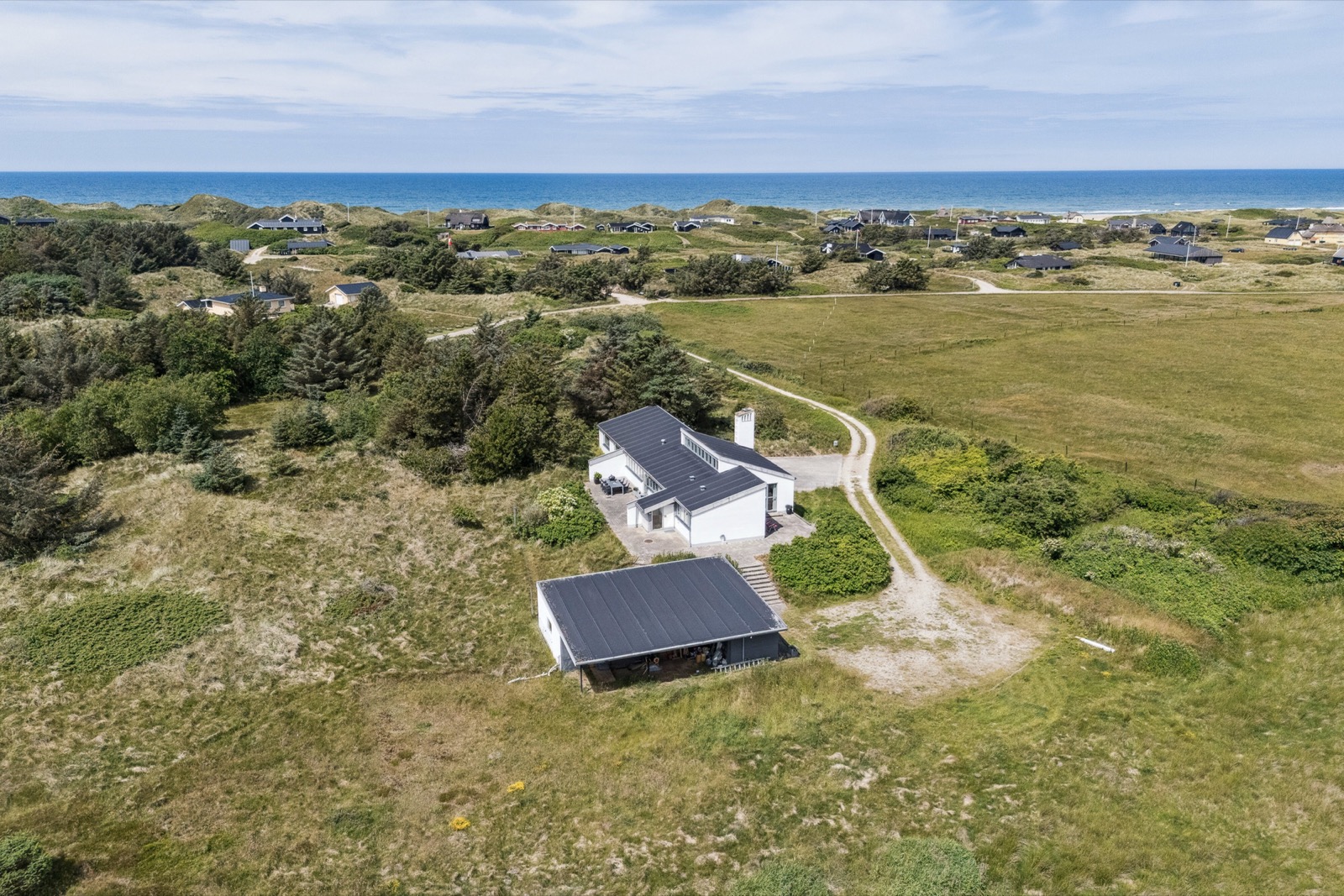 Dronefoto af ejendom og grund med det hvide hus, sorttaget garage, indkørsel og klitlandskab mod havet.